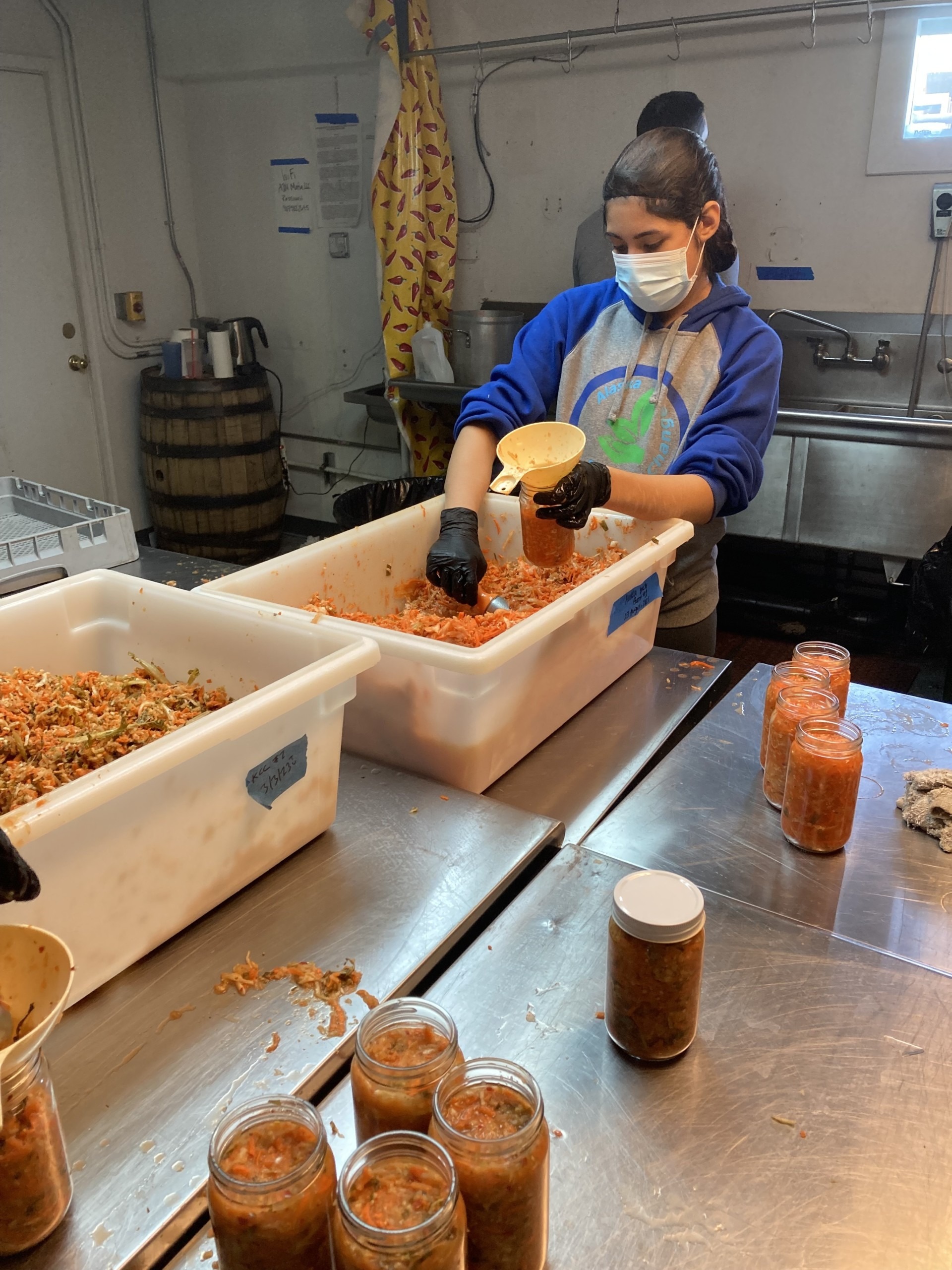 woman in mask/hairnet, gloves ladling kimchi into jars